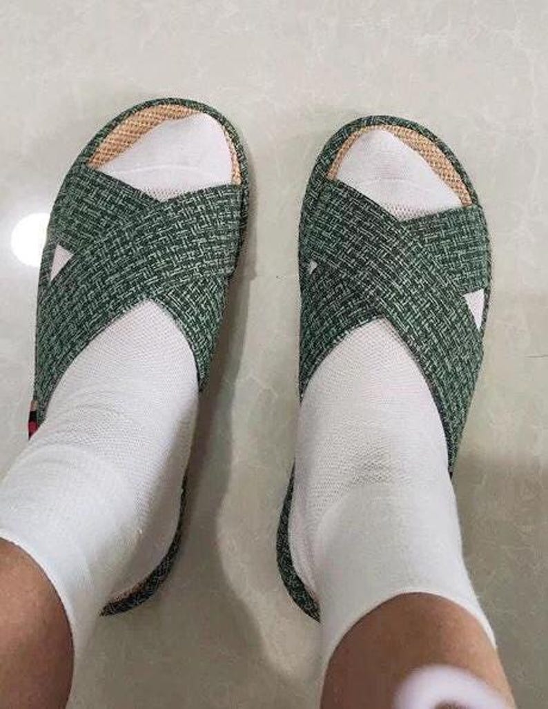 K&ouml;nnte beinhalten: Ein Paar gr&uuml;n gewebte Sandalen mit Kreuzmuster, getragen mit wei&szlig;en Socken. Die Sandalen haben eine naturfarbene Einfassung und einen schlichten, l&auml;ssigen Stil, perfekt f&uuml;r den Innen- oder Au&szlig;enbereich.