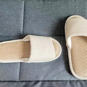 May include: Two beige and tan indoor slippers. The footbed is woven, and the top strap is a solid beige fabric. The slippers are placed on a gray textured surface.