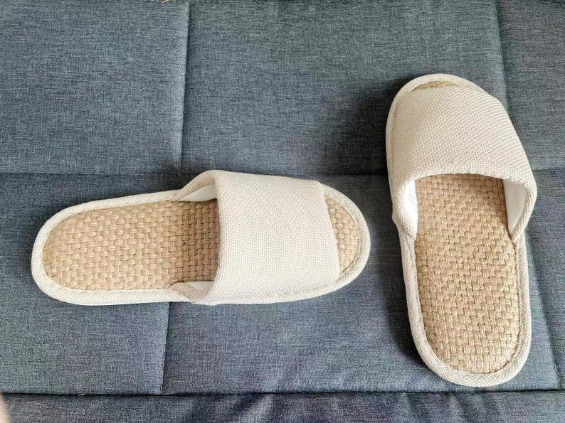 May include: Two beige and tan indoor slippers. The footbed is woven, and the top strap is a solid beige fabric. The slippers are placed on a gray textured surface.