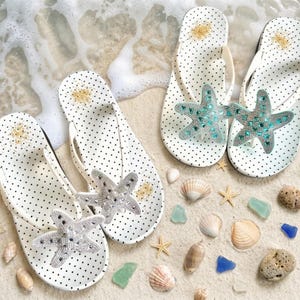May include: White polka dot flip-flops with a starfish embellishment. The flip-flops have white straps and are decorated with a sparkling, bejeweled starfish. The flip-flops are on a sandy surface with seashells and sea glass.