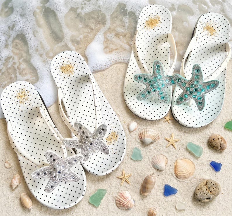 May include: White polka dot flip-flops with a starfish embellishment. The flip-flops have white straps and are decorated with a sparkling, bejeweled starfish. The flip-flops are on a sandy surface with seashells and sea glass.