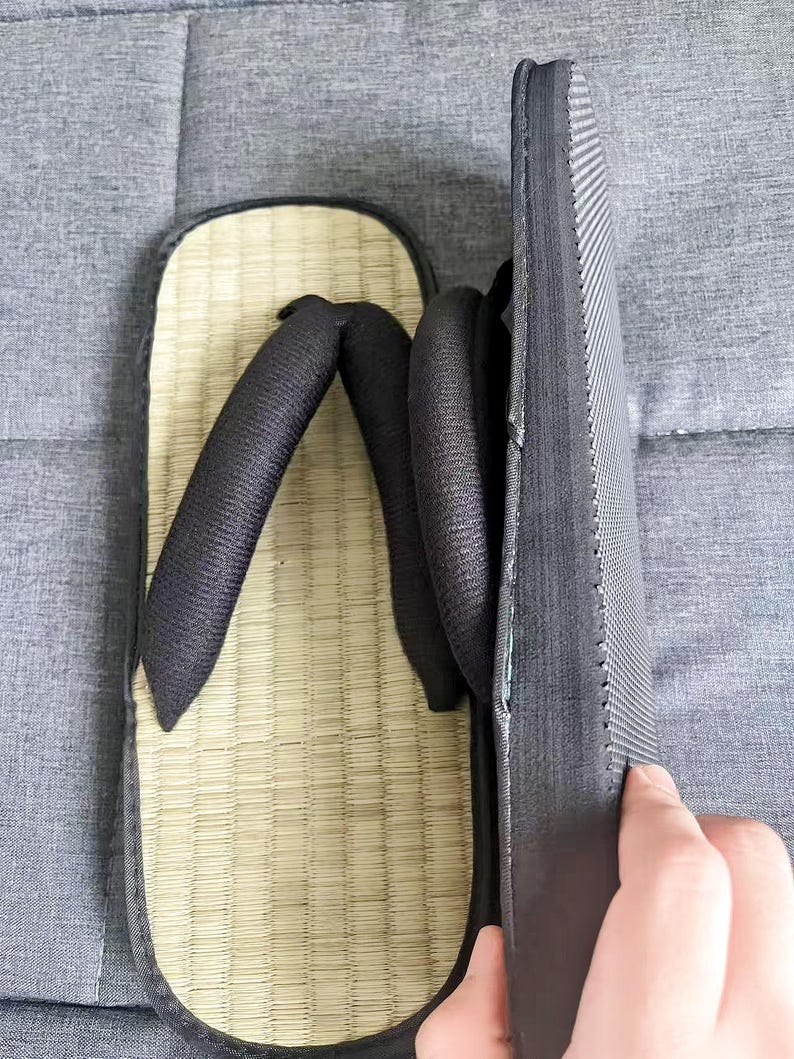 May include: A pair of traditional Japanese geta sandals. The sandals feature a natural woven straw footbed, black straps, and a black sole. The geta are designed with a raised platform for comfort and are suitable for casual wear.