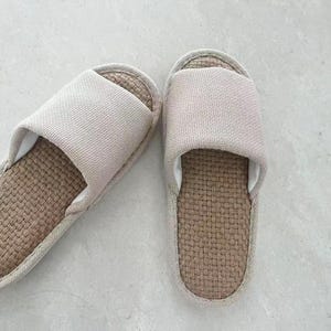 May include: A pair of beige linen slippers. The top strap is a solid beige color, and the footbed is a woven brown material. The slippers are designed for indoor use and provide comfort and breathability.