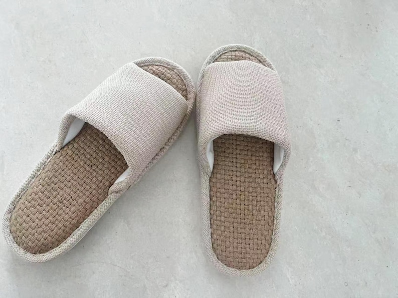 May include: A pair of beige linen slippers. The top strap is a solid beige color, and the footbed is a woven brown material. The slippers are designed for indoor use and provide comfort and breathability.