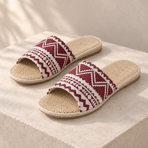 May include: A pair of open-toe slide sandals featuring a woven tan jute footbed and a wide fabric strap across the top. The strap displays a deep burgundy and white geometric pattern with repeating zigzag and diamond shapes throughout the design.