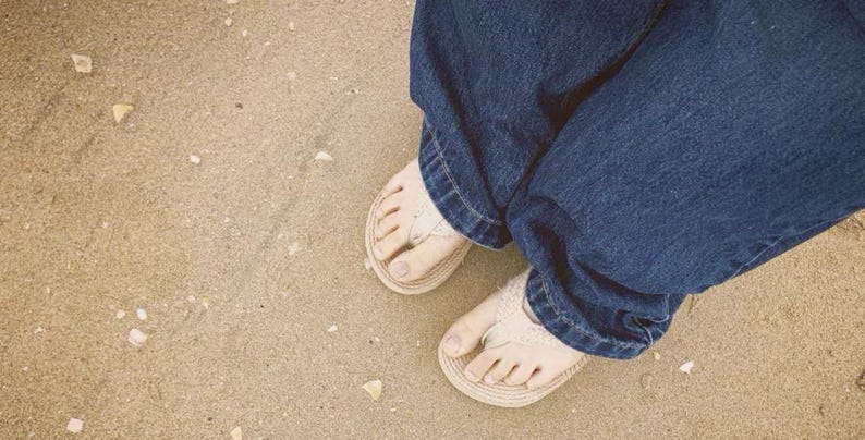 Może przedstawiać: Osoba w niebieskich dżinsach i beżowych sandałach z białymi paskami, odpoczywająca na piaszczystej plaży. Zdjęcie zrobione z g&oacute;ry, pokazujące stopy i dolne nogi.