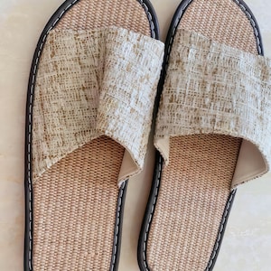 May include: A pair of open-toe slippers featuring a woven tan footbed and a textured beige and white fabric upper strap. The slippers are finished with a dark brown trim and white stitching along the outer edge of the sole for a clean look.