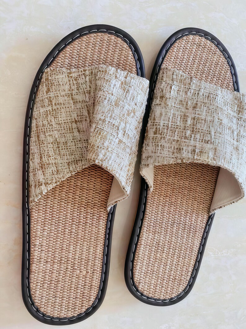 May include: A pair of open-toe slippers featuring a woven tan footbed and a textured beige and white fabric upper strap. The slippers are finished with a dark brown trim and white stitching along the outer edge of the sole for a clean look.