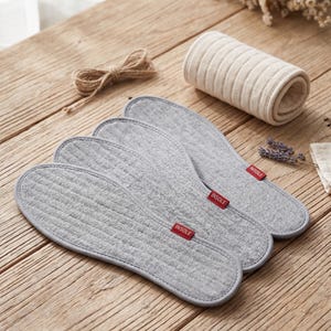 Può includere: Tre solette in tessuto grigio con etichette rosse "INSOLE" sono disposte su una superficie di legno. Sono presenti anche un oggetto di tessuto beige arrotolato e uno spago. Le solette hanno una trama trapuntata.