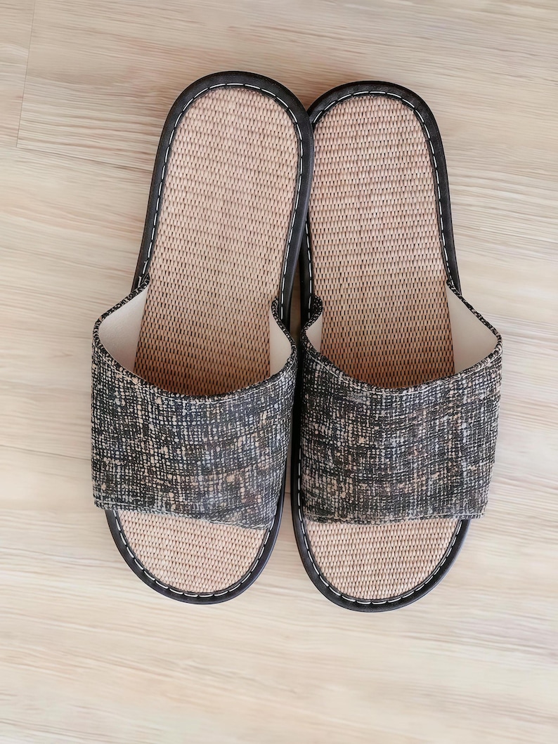 May include: A pair of open-toe slippers featuring a textured, dark charcoal and tan woven fabric upper. The footbed is made of a light beige, woven material with a smooth, dark brown trim outlining the entire edge of each slipper.