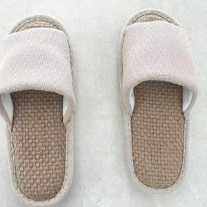 May include: A pair of beige linen slippers. Each slipper features a woven, light brown footbed and a matching beige fabric upper. The interior is white. The slippers are designed for indoor use.