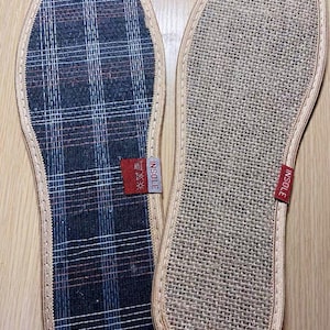May include: Two shoe insoles. One insole has a navy blue, red, and white plaid pattern. The other insole is a beige woven material. Both insoles have a beige border and a red tag that says "INSOLE".