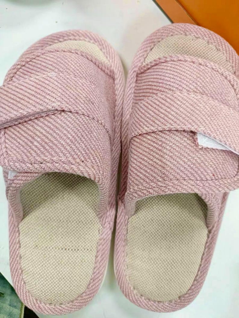 May include: A pair of pink fabric slippers with a striped pattern. The slippers have a beige insole and an adjustable strap. The design is simple and comfortable, suitable for indoor wear.
