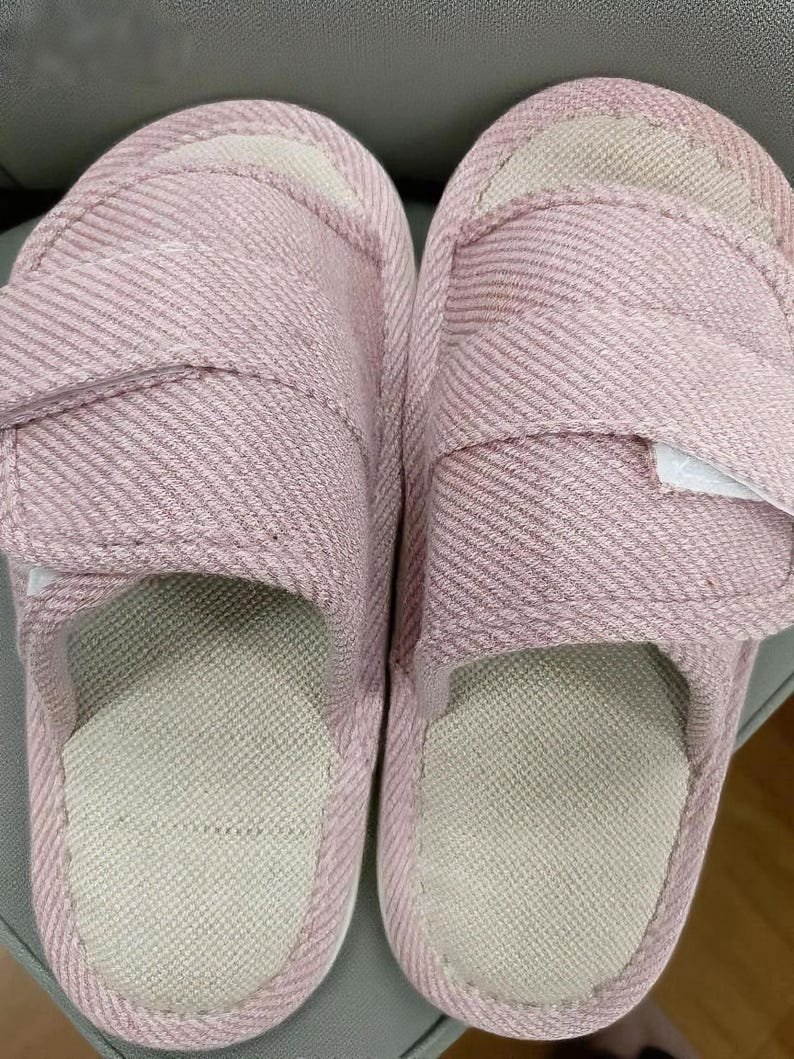 May include: A pair of pink adjustable slippers with a textured fabric. The slippers have a light beige interior and a hook-and-loop closure for a secure fit. The sole is a light beige color.