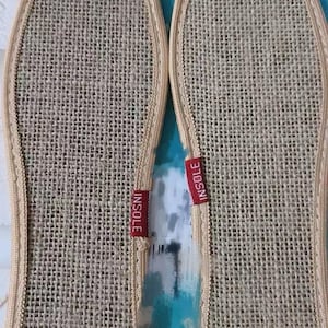 May include: Two beige woven insoles with a stitched border. Each insole has a red tag that reads "INSOLE". The insoles are designed to fit the shape of a foot and are likely for shoes or slippers.