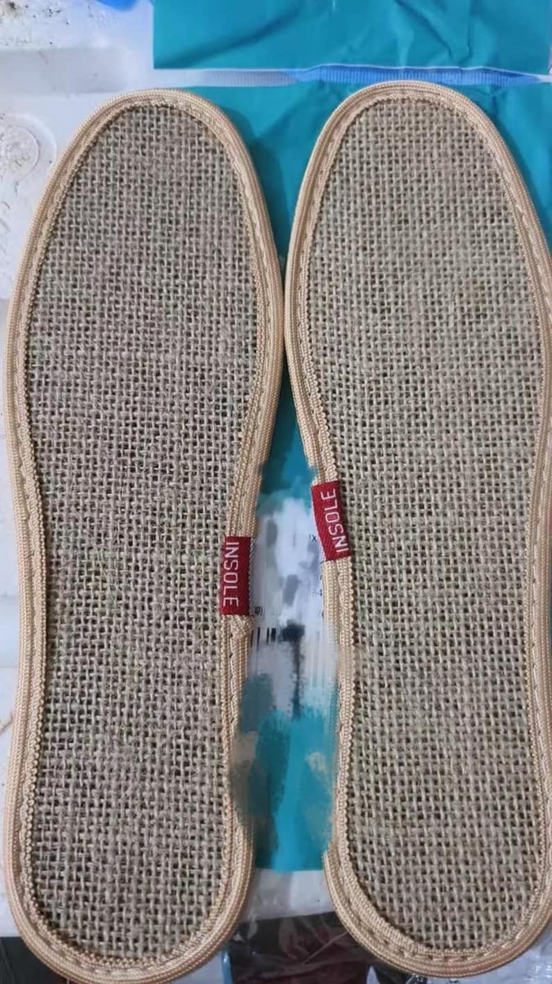 May include: Two beige woven insoles with a stitched border. Each insole has a red tag that reads "INSOLE". The insoles are designed to fit the shape of a foot and are likely for shoes or slippers.