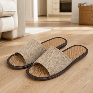 May include: A pair of open-toe slippers featuring a beige fabric upper strap and a woven tan footbed. The slippers are finished with a dark brown trim around the edges of the sole, providing a structured look for indoor footwear use.