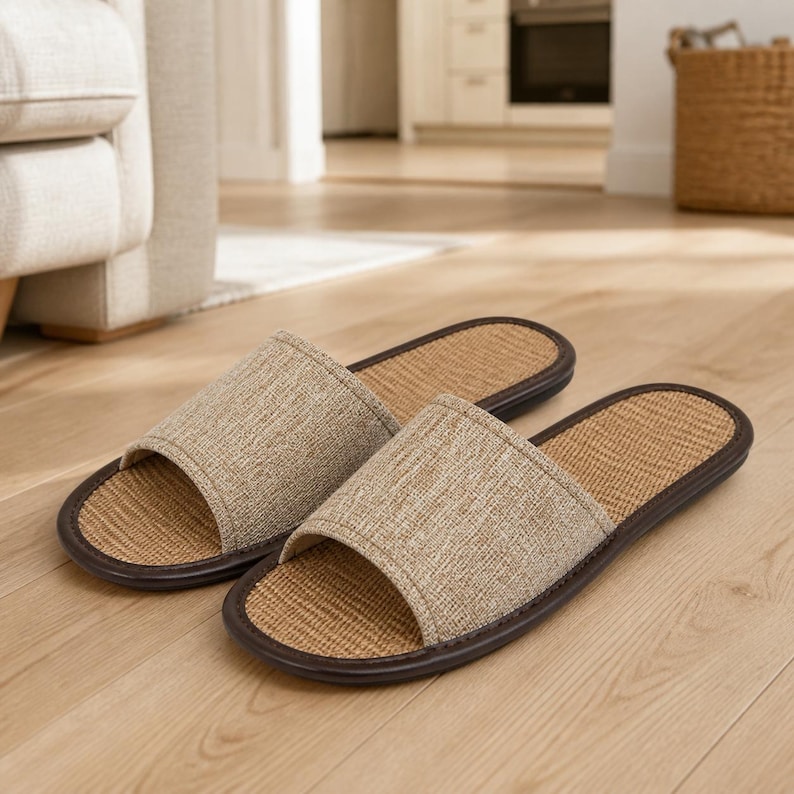 May include: A pair of open-toe slippers featuring a beige fabric upper strap and a woven tan footbed. The slippers are finished with a dark brown trim around the edges of the sole, providing a structured look for indoor footwear use.