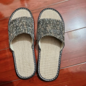 May include: A pair of open-toe slippers featuring a textured beige woven footbed and a dark gray and tan mottled fabric upper strap. The slippers are finished with a dark brown trim and white stitching along the edges of the sole and strap.