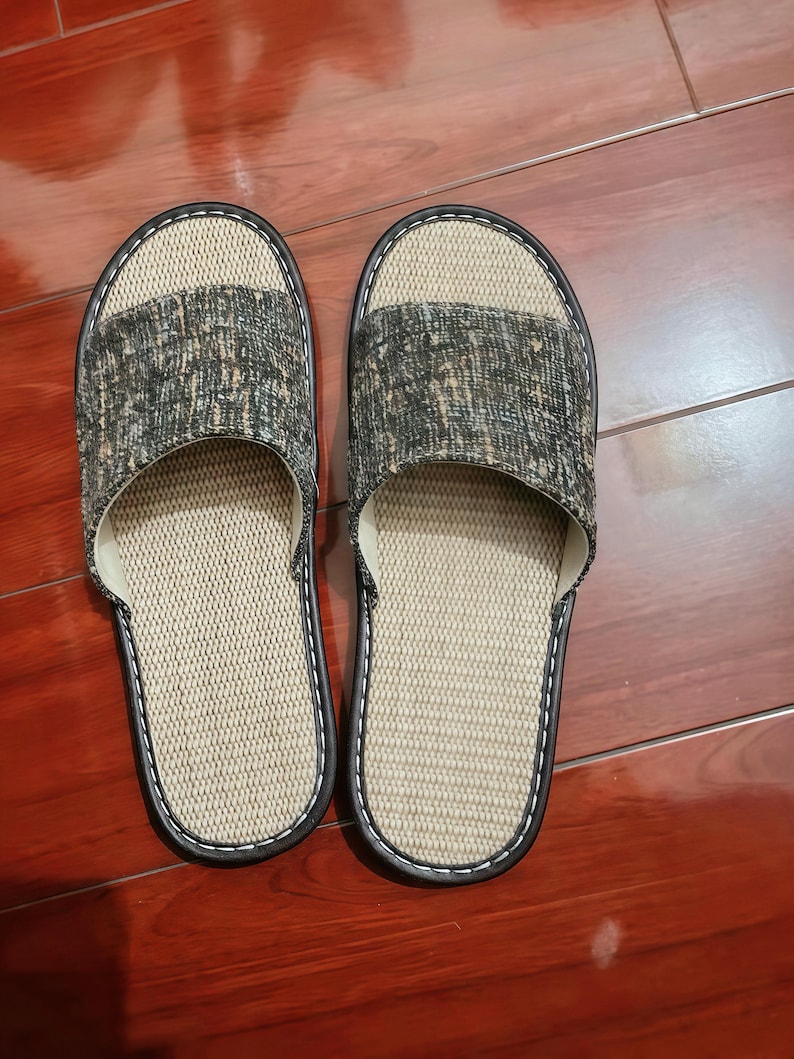 May include: A pair of open-toe slippers featuring a textured beige woven footbed and a dark gray and tan mottled fabric upper strap. The slippers are finished with a dark brown trim and white stitching along the edges of the sole and strap.