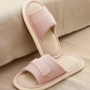 May include: Two pink fabric slippers with open toes and a strap across the top. The slippers have a light beige interior and a white sole. The slippers are resting on a beige surface.