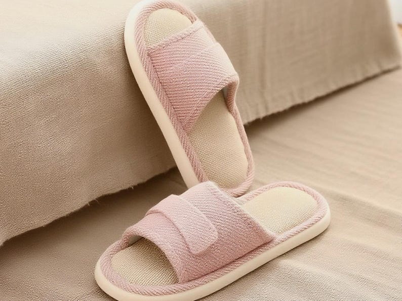 May include: Two pink fabric slippers with open toes and a strap across the top. The slippers have a light beige interior and a white sole. The slippers are resting on a beige surface.