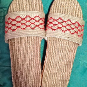 May include: A pair of beige woven slide sandals. The sandals have a textured footbed and a wide strap across the top. The strap is decorated with a red geometric pattern. The sandals are made of natural materials and appear to be handmade.