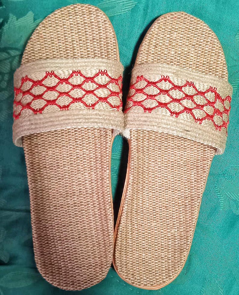 May include: A pair of beige woven slide sandals. The sandals have a textured footbed and a wide strap across the top. The strap is decorated with a red geometric pattern. The sandals are made of natural materials and appear to be handmade.