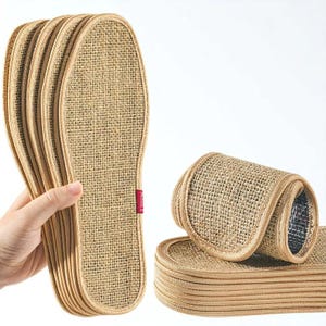 May include: A stack of woven insoles made of natural fibers, with a light brown color. The insoles are oval-shaped and have a textured surface. A few insoles are rolled up, showing a silver lining. The word "INSOLE" is printed on a red tag.