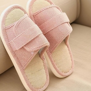 May include: A pair of pink fabric slippers with a light beige interior and white soles. The slippers have a strap across the top and are designed for indoor use. The slippers are made of a soft, textured material.