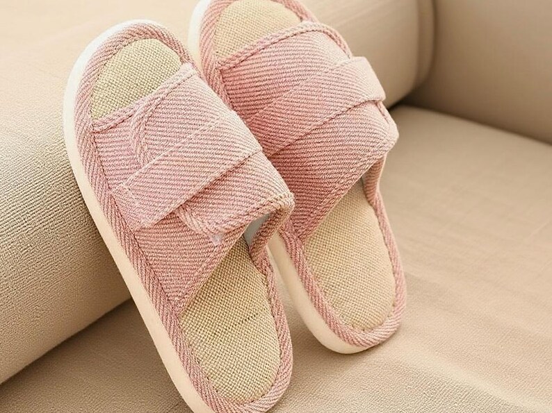 May include: A pair of pink fabric slippers with a light beige interior and white soles. The slippers have a strap across the top and are designed for indoor use. The slippers are made of a soft, textured material.