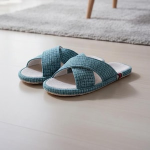 May include: A pair of teal cross-strap slippers. The slippers have a textured fabric upper and a white inner sole. The soles are a light beige color with a teal border. The slippers are on a light wood floor.
