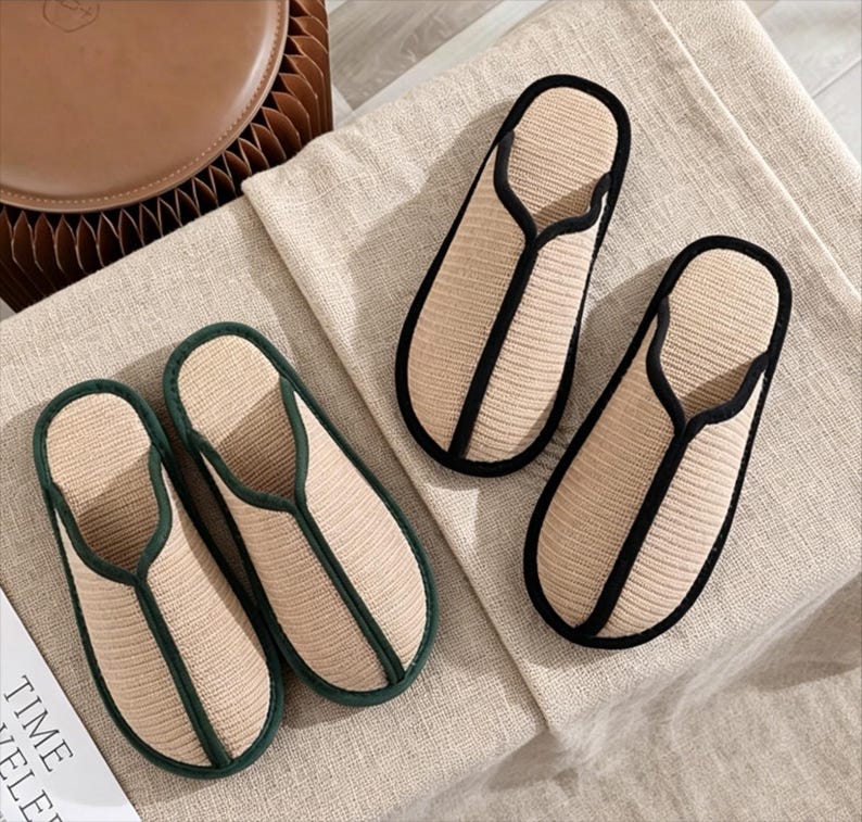 Pu&ograve; includere: Due paia di pantofole beige e nere con una trama intrecciata. Un paio ha un bordo verde scuro. Le pantofole sono disposte su un tessuto di colore neutro, suggerendo un focus sul comfort e l'uso domestico.