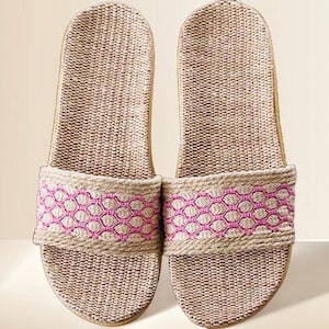 May include: A pair of woven beige and tan slide sandals. The sandals feature a pink geometric pattern across the top strap. The soles are a light tan color. These casual sandals are perfect for indoor or outdoor wear.