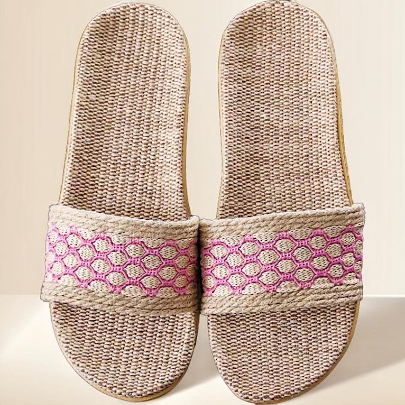 May include: A pair of woven beige and tan slide sandals. The sandals feature a pink geometric pattern across the top strap. The soles are a light tan color. These casual sandals are perfect for indoor or outdoor wear.