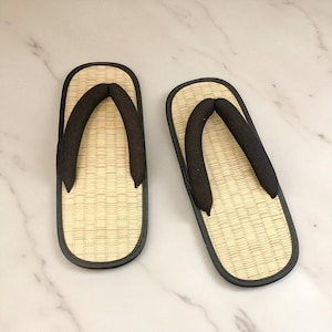 May include: A pair of traditional Japanese geta sandals. The sandals feature a woven straw footbed, a black thong strap, and a black border. The geta are designed for outdoor wear and are a common type of footwear in Japan.