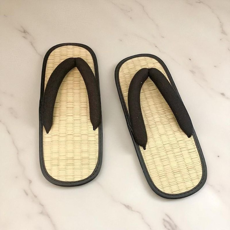 May include: A pair of traditional Japanese geta sandals. The sandals feature a woven straw footbed, a black thong strap, and a black border. The geta are designed for outdoor wear and are a common type of footwear in Japan.
