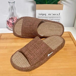 Könnte beinhalten: Ein Paar braune und beige gewebte Hausschuhe. Die Hausschuhe haben ein strukturiertes Design mit offener Spitze, ein braunes Obermaterial und ein beiges Fußbett. Die Hausschuhe bestehen aus Naturfasermaterial und sind für den Innenbereich geeignet.