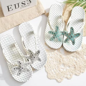 May include: White polka dot flip-flops with a starfish embellishment. The flip-flops have a white strap and a decorative starfish made of silver and turquoise-colored rhinestones. The footwear is displayed on a white surface.