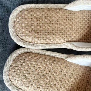 May include: Close-up of two beige woven slippers. The slippers have a textured, woven design on the footbed and a light-colored trim around the edges. The interior appears to be a soft, white fabric. The slippers are likely for indoor use.
