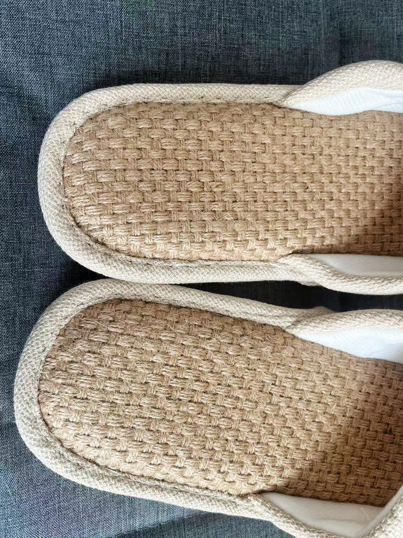 May include: Close-up of two beige woven slippers. The slippers have a textured, woven design on the footbed and a light-colored trim around the edges. The interior appears to be a soft, white fabric. The slippers are likely for indoor use.