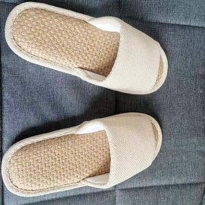 May include: A pair of beige open-toe slippers. The footbed is made of woven natural fibers, and the upper is a solid, light-colored fabric. The slippers are designed for indoor use and rest on a gray surface.