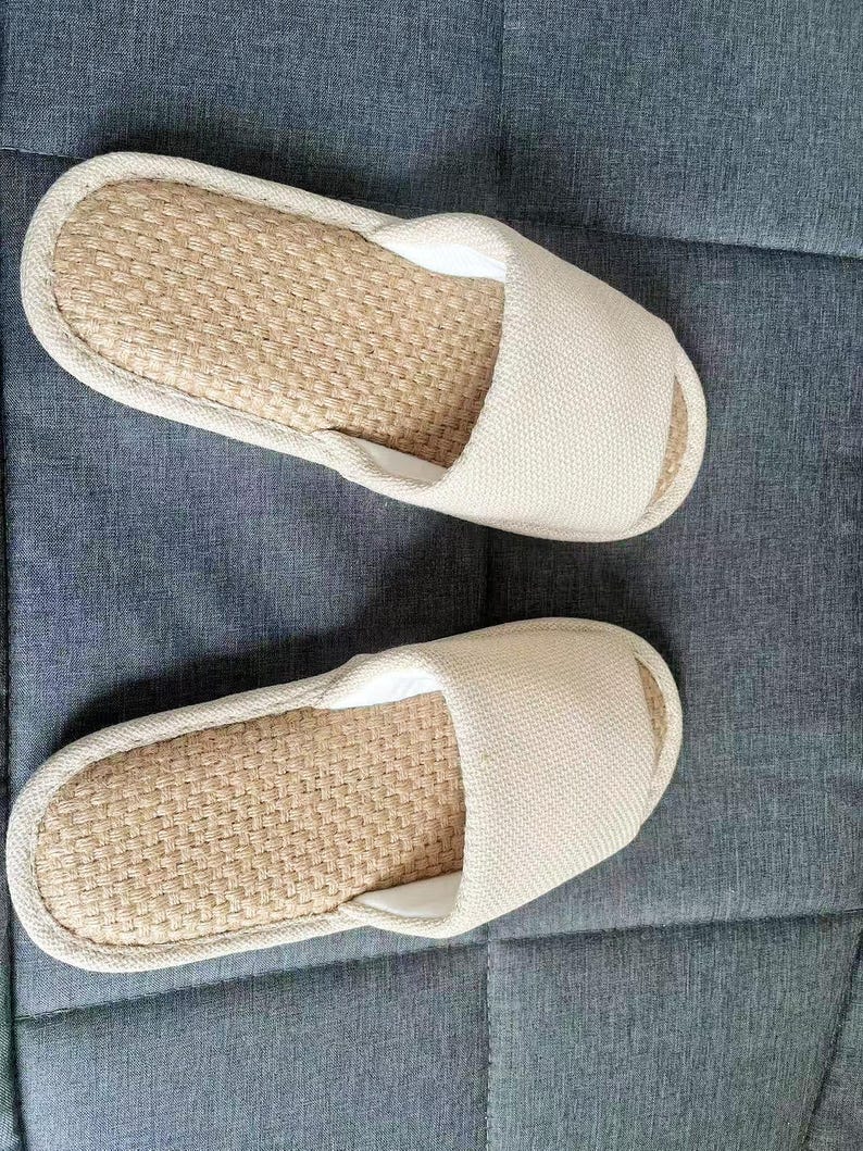 May include: A pair of beige open-toe slippers. The footbed is made of woven natural fibers, and the upper is a solid, light-colored fabric. The slippers are designed for indoor use and rest on a gray surface.