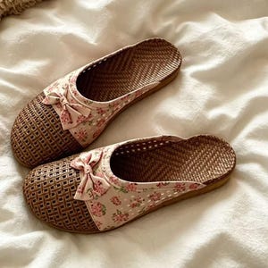 May include: A pair of woven brown slippers with a floral print and bow detail. The slippers have a light pink fabric with a small pink floral pattern. The soles are a light brown colour.