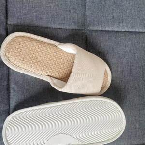 May include: Pair of beige open-toe slippers. The top of the slipper has a woven, natural fiber footbed and a beige fabric strap. The sole is white with a wavy pattern for traction. The slippers are on a gray surface.