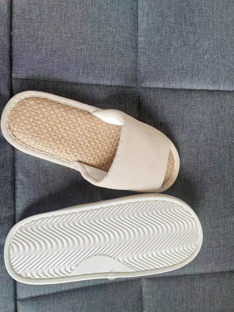 May include: Pair of beige open-toe slippers. The top of the slipper has a woven, natural fiber footbed and a beige fabric strap. The sole is white with a wavy pattern for traction. The slippers are on a gray surface.