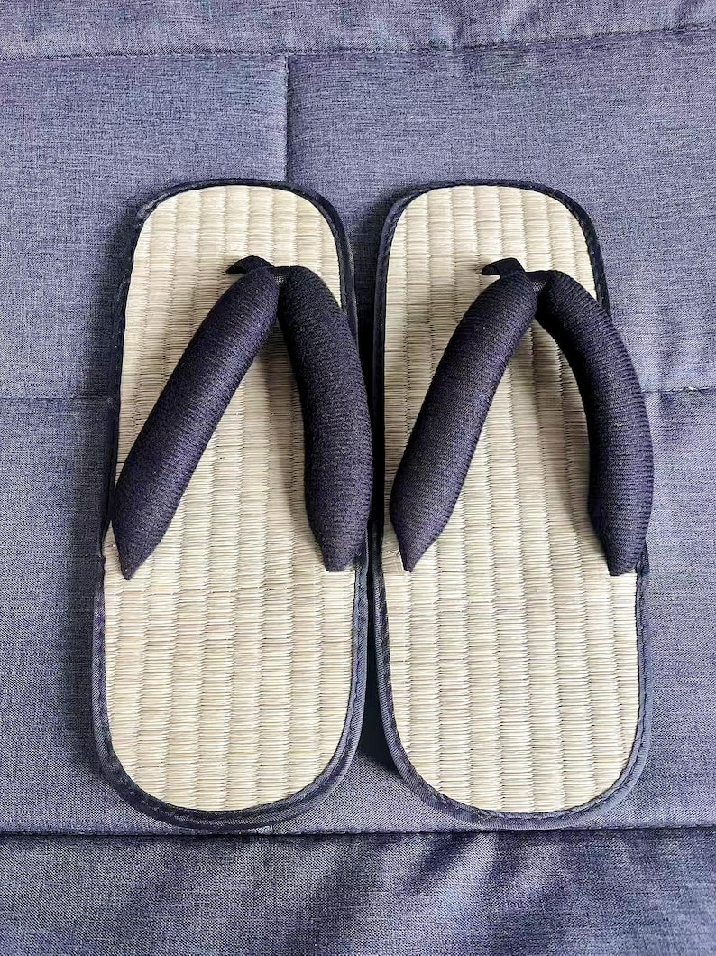 May include: A pair of traditional Japanese geta sandals. The sandals feature a woven straw footbed, a dark blue border, and dark blue fabric straps. The geta are placed on a blue fabric surface.