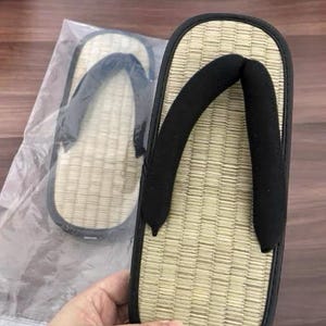 May include: Tan and black Japanese geta sandals. The sandals have a woven straw footbed, a black strap, and a black rubber sole. One sandal is held in a hand, and another is in a plastic bag.