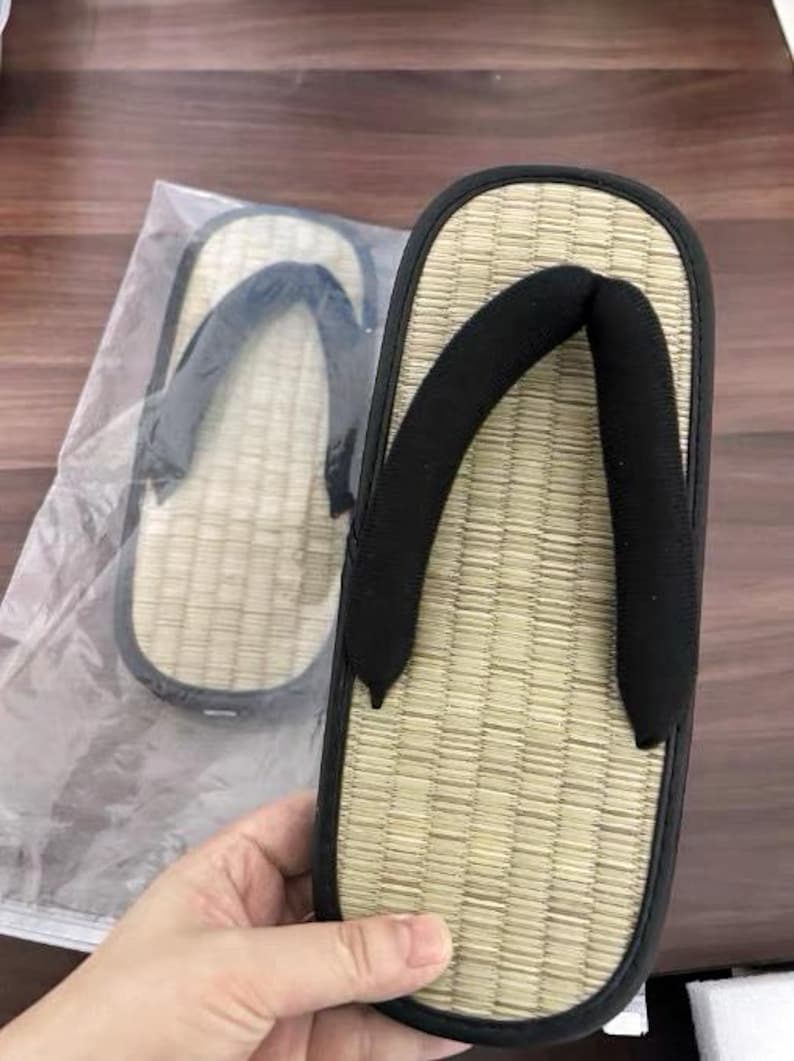 May include: Tan and black Japanese geta sandals. The sandals have a woven straw footbed, a black strap, and a black rubber sole. One sandal is held in a hand, and another is in a plastic bag.