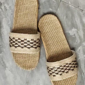 May include: A pair of beige woven sandals. The sandals have a textured, open-toe design with a decorative band featuring a repeating pattern in a darker brown color. The soles appear to be made of a light-colored wood or similar material.